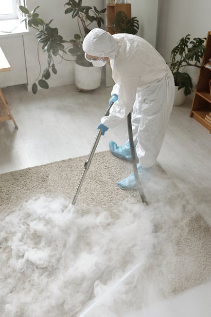 A person dressed in full cleaning protective gear, including a white coverall, face mask, gloves, and shoe covers, is performing deep carpet cleaning on a beige rug using a steam cleaning machine with