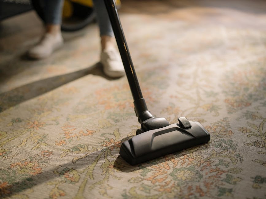 A person dressed in full cleaning protective gear, including a white coverall, face mask, gloves, and shoe covers, is performing deep carpet cleaning on a beige rug using a steam cleaning machine with