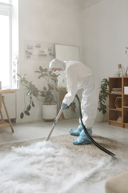 A person dressed in grey work overalls is using a floor squeegee to clean a dark grey carpeted surface in a dimly lit indoor space, possibly a commercial or conference area. The individual is bending