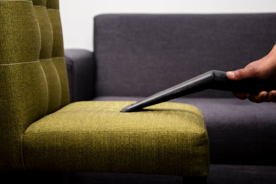 A close-up of a person's hand holding a handheld steam cleaner nozzle, applying deep cleaning to the beige fabric upholstery of a car seat. The upholstery has a textured pattern with darker stains or