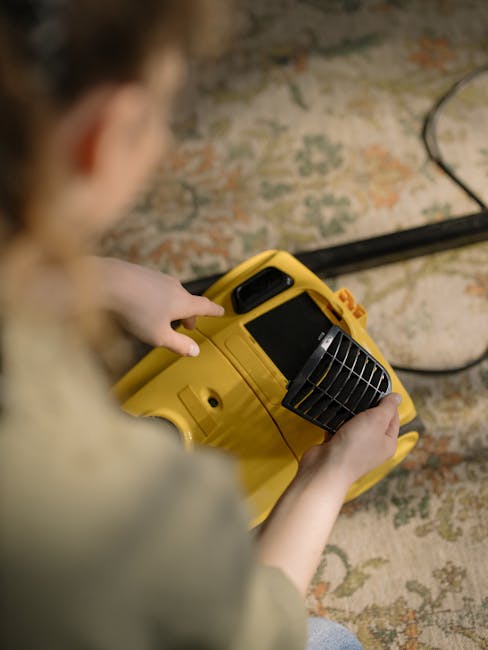 A person holding a yellow portable industrial fan or dehumidifier in a room with a patterned carpet, possibly during a cleaning or drying process. The carpet features muted tones of beige, green, and orange with intricate floral and geometric designs. The scene shows the individual positioned near a wall or corner, with the fan directed towards a surface for surface cleaning, sanitisation, or moisture removal as part of a professional cleaning service. Visible are the person's hands, a portion of their arm, and their blurred head, emphasizing the focus on the cleaning equipment. The room is well-lit, highlighting the cleanliness and organized setting, with Kingston Vale Carpet Cleaners' service potentially involved in maintaining hygiene and surface cleanliness according to the page content.