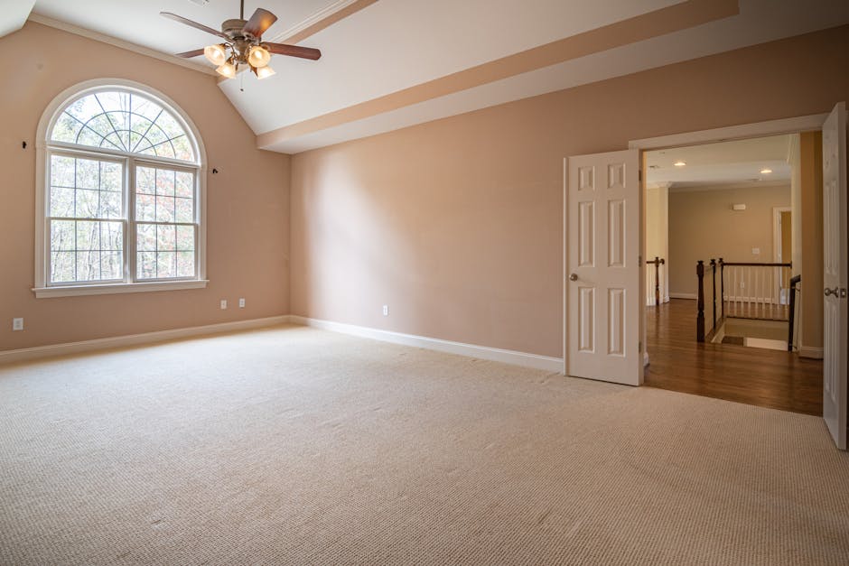 A spacious, empty room with beige walls and a large arched window letting in natural light. The room features a plush, light-colored carpet, a ceiling fan with attached light fixtures, and a white door open to an adjacent area with hardwood flooring and a staircase railing. The space appears clean and well-maintained, reflecting comprehensive surface cleaning and domestic sanitisation possibly provided by Kingston Vale Carpet Cleaners, as suggested by the context of the webpage about the real cost of carpet cleaning in Kingston Vale.