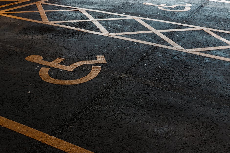 Two blue wheelchair-accessible parking signs mounted on yellow posts along a concrete sidewalk in Kingston Vale, with several parked cars, including a maroon SUV and a black vehicle, visible in the ba